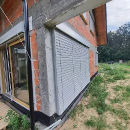 Szare rolety zewnętrzne zamontowane na surowej elewacji domu w trakcie budowy, widoczne fragmenty cegieł, betonu i instalacji.