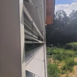 Detaliczny widok z boku na częściowo otwarte, białe żaluzje fasadowe z widocznym mechanizmem sterującym, na tle rozmytego ogrodu i fragmentu nieba.