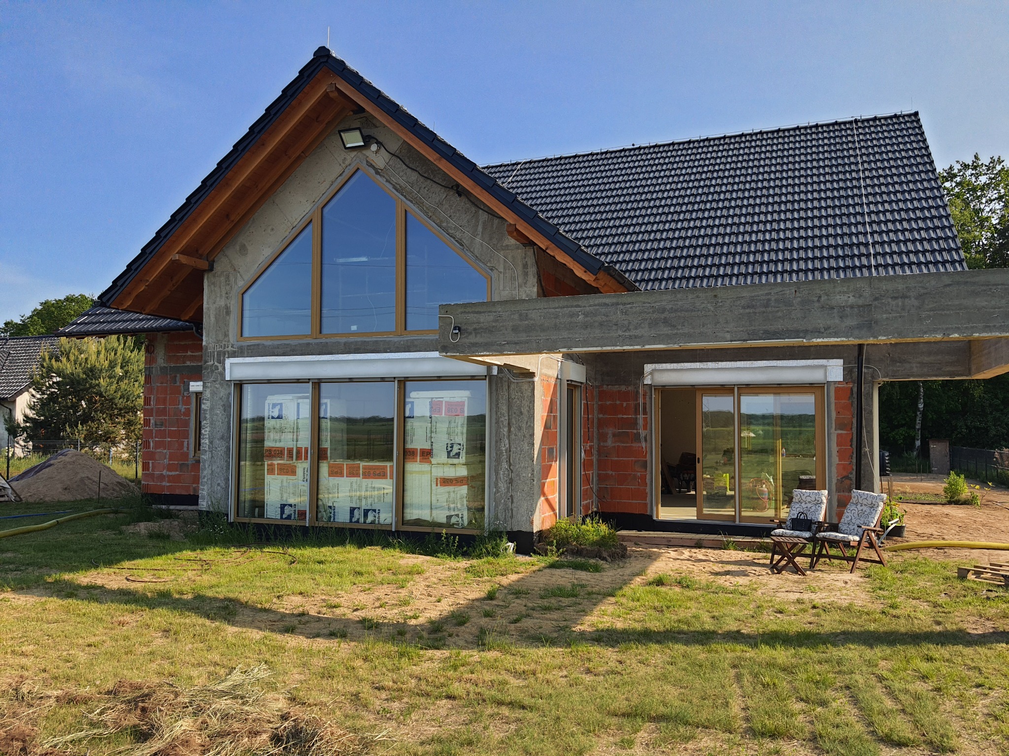 Surowy stan domu jednorodzinnego z dużymi oknami PCV, częściowo zakrytymi folią ochronną, z widocznymi elementami konstrukcyjnymi i przygotowanym terenem wokół.