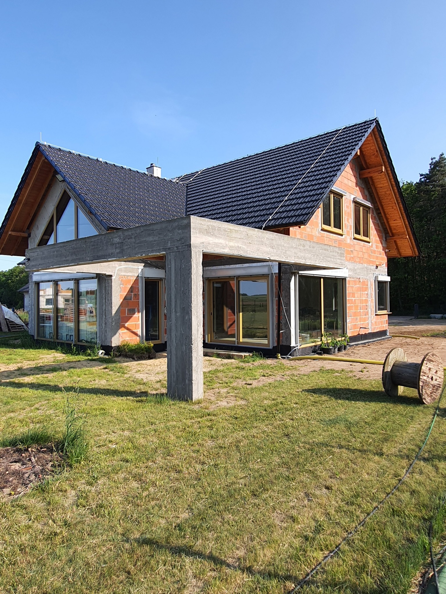 Dom w trakcie budowy z widocznymi oknami z ramami w kolorze drewna, surowymi ścianami z czerwonej cegły i betonową konstrukcją werandy, na trawniku leży drewniana szpula.