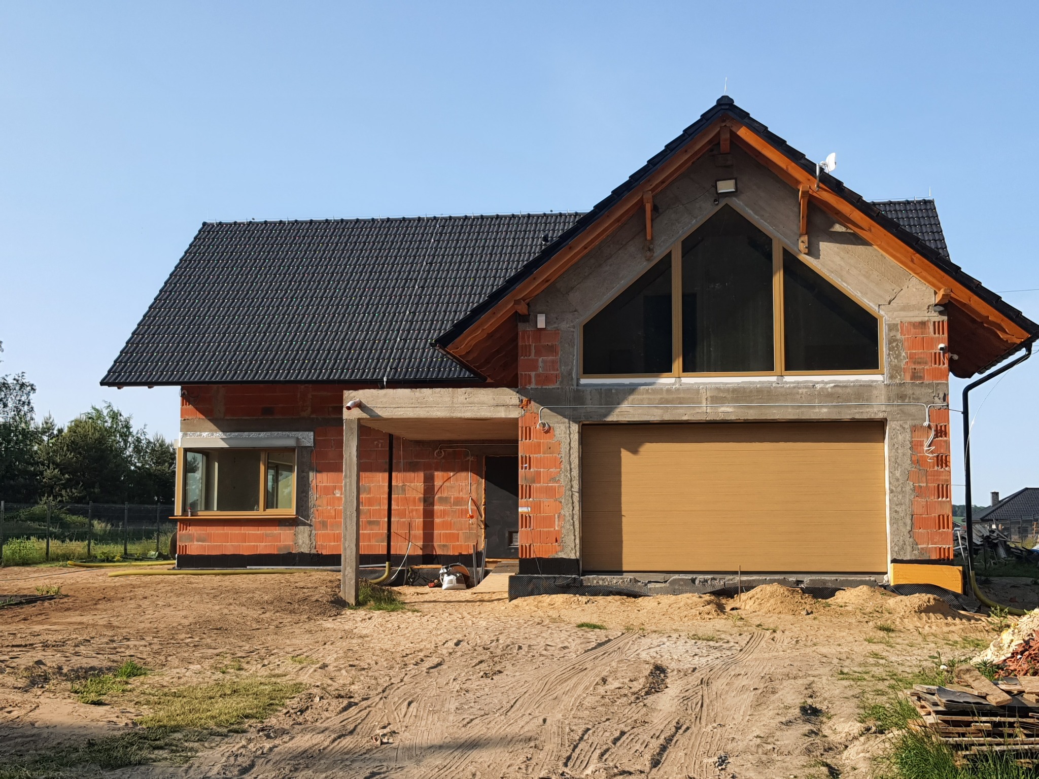 Niedokończony dom jednorodzinny z zamontowaną segmentową bramą garażową w kolorze piaskowym, widoczne okna z drewnianą ramą, ściany z czerwonej cegły i czarny dach z dachówki ceramicznej.