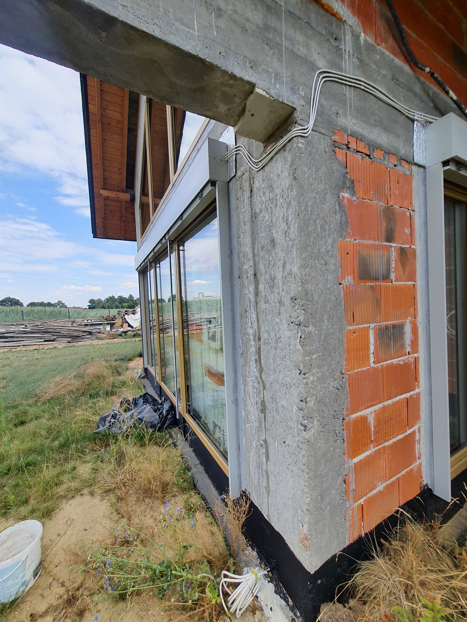 Surowy stan budowy domu z zamontowanymi oknami z drewnianą ramą, widoczne niezabezpieczone kable elektryczne, fragmenty muru z cegły i betonu oraz składowane deski w tle.