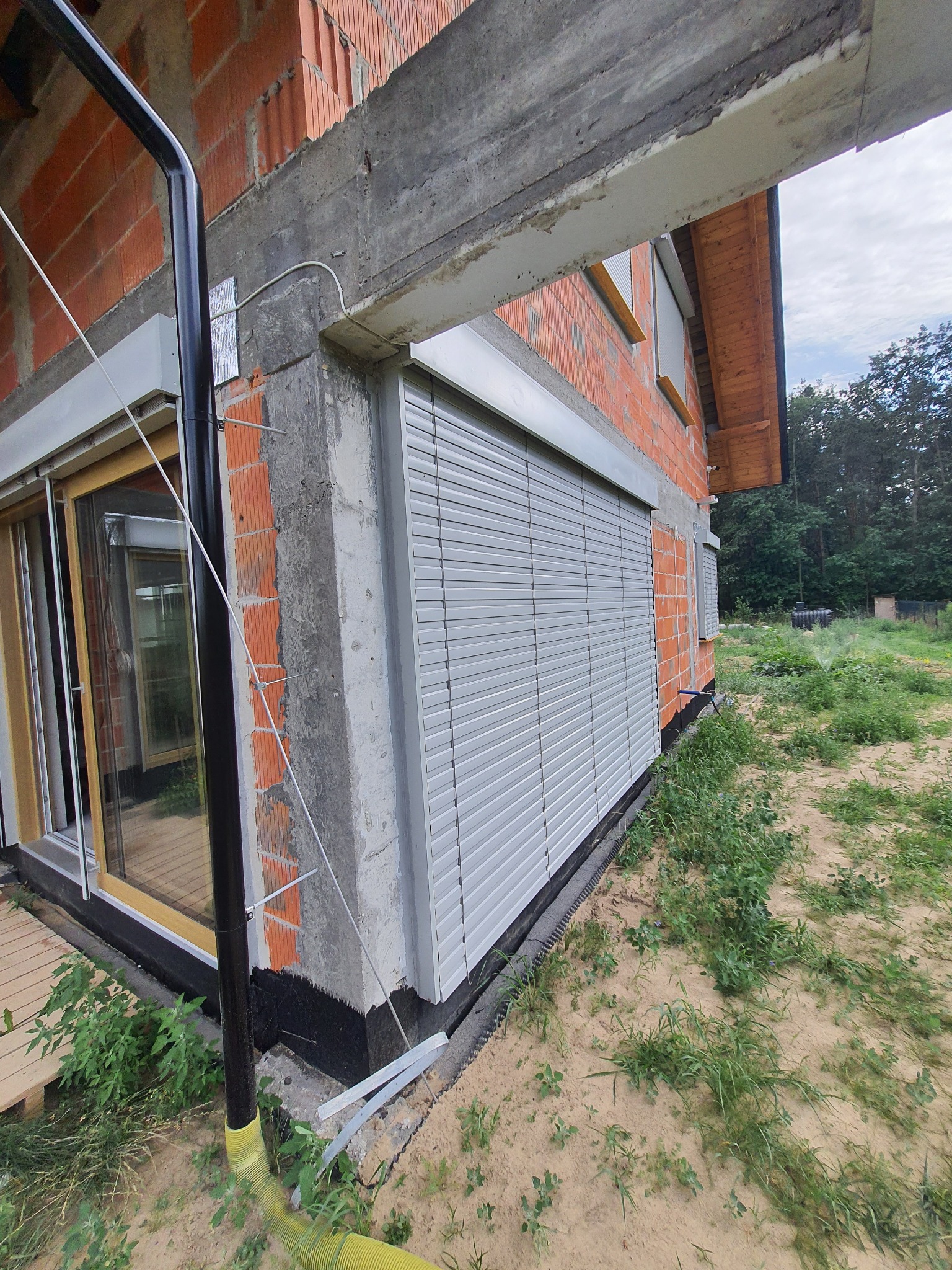 Szare rolety zewnętrzne zamontowane na surowej elewacji domu w trakcie budowy, widoczne fragmenty cegieł, betonu i instalacji.