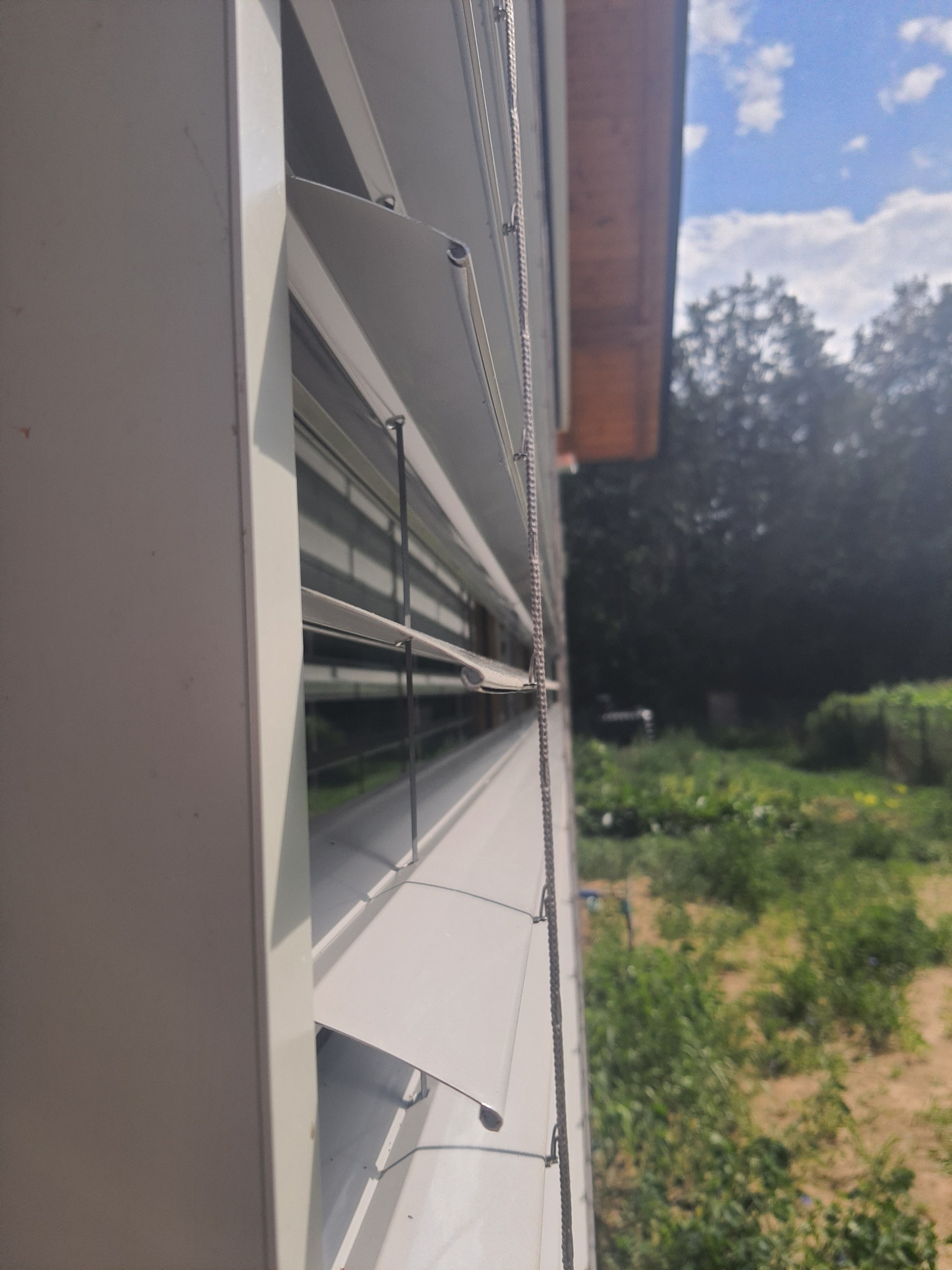 Detaliczny widok z boku na częściowo otwarte, białe żaluzje fasadowe z widocznym mechanizmem sterującym, na tle rozmytego ogrodu i fragmentu nieba.