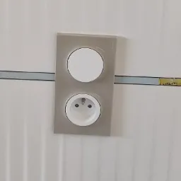 Szary, podwójny panel z gniazdkiem elektrycznym i wyłącznikiem, zamontowany na białej ścianie z pionowymi paskami, widoczna szara listwa dekoracyjna.