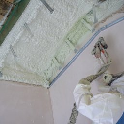 TOMCZAK - IZOLACJE - Poddasze ocieplone pianką PUR z widocznymi mocowaniami i aplikatorem. Praca w toku, pianka świeżo nałożona, widoczne detale struktury i sprzęt ochronny.