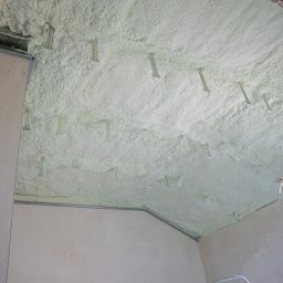 TOMCZAK - IZOLACJE - Poddasze po aplikacji pianki PUR, widoczne mocowania. Izolacja pokrywa całą powierzchnię, tworząc jednolitą warstwę. W tle ściana i drabina.