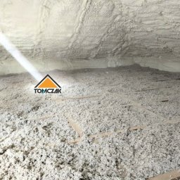 TOMCZAK - IZOLACJE - Poddasze po ociepleniu pianką PUR, widoczne drewniane belki konstrukcyjne, nierównomierna struktura pianki, logo firmy w lewym dolnym rogu.