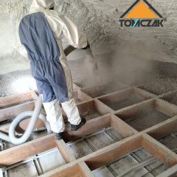 TOMCZAK - IZOLACJE - Pracownik w kombinezonie aplikuje pianę PUR na poddaszu. Widoczne drewniane legary i warstwa izolacji. Logo firmy w prawym górnym rogu.