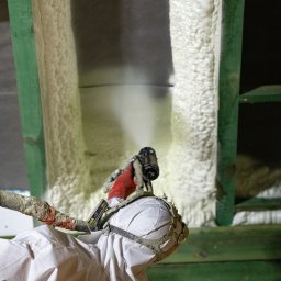 TOMCZAK - IZOLACJE - Pracownik w kombinezonie nanosi pianę PUR na drewnianą konstrukcję, tworząc izolację termiczną. Widoczny pistolet natryskowy i fragment ocieplonej powierzchni.