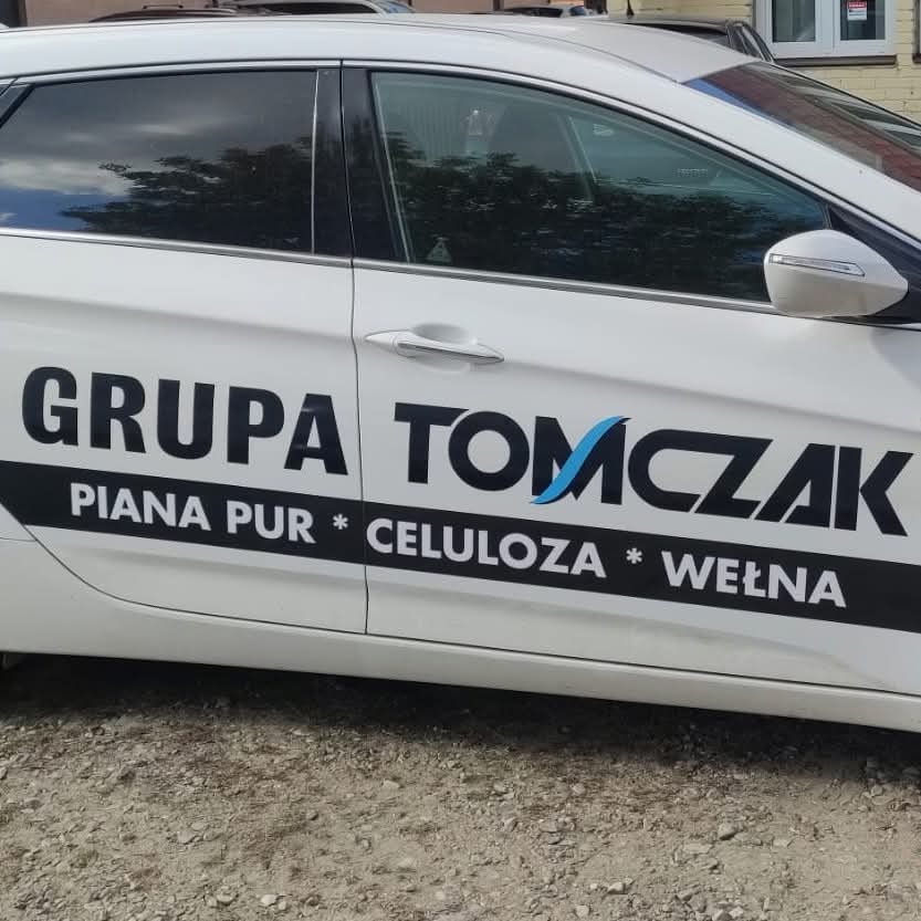 Biały samochód z logo firmy 'Grupa Tomczak', specjalizującej się w ocieplaniu pianą PUR, celulozą i wełną, zaparkowany na żwirowym podłożu.
