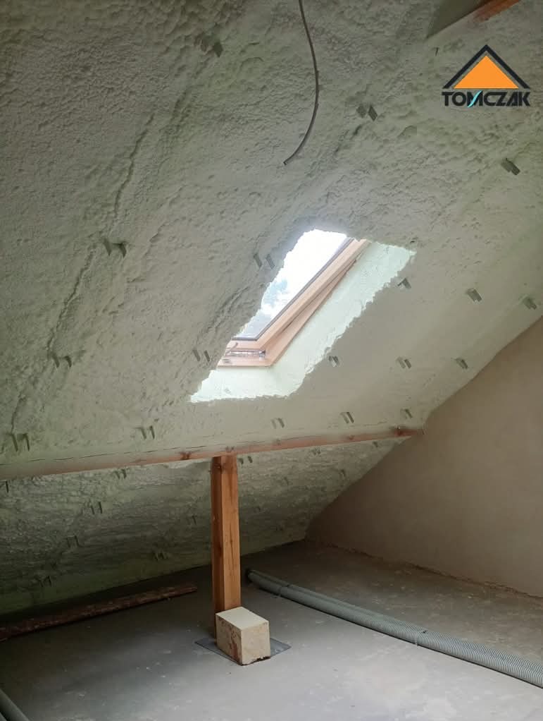 Poddasze po ociepleniu pianką PUR, widoczne krokwie i okno dachowe. Biała pianka pokrywa całą powierzchnię, tworząc jednolitą izolację. Na podłodze szary przewód wentylacyjny.