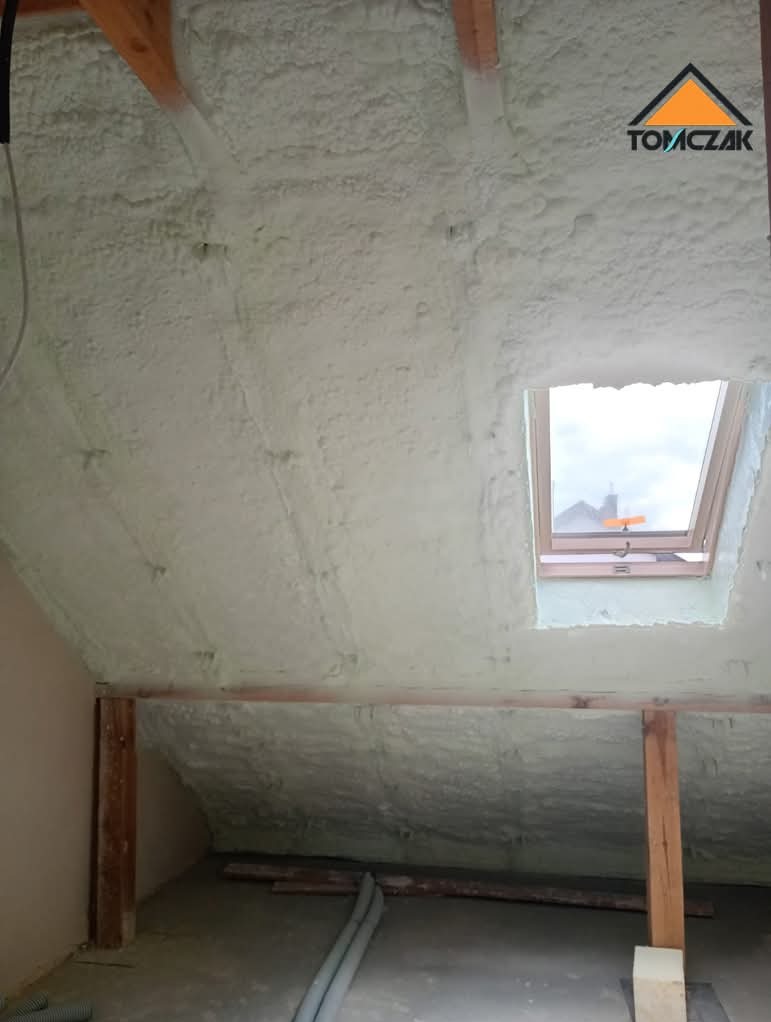 Poddasze po ociepleniu pianką PUR, widoczne drewniane elementy konstrukcyjne i okno dachowe. Biała, nierówna powierzchnia pianki pokrywa skośne ściany.