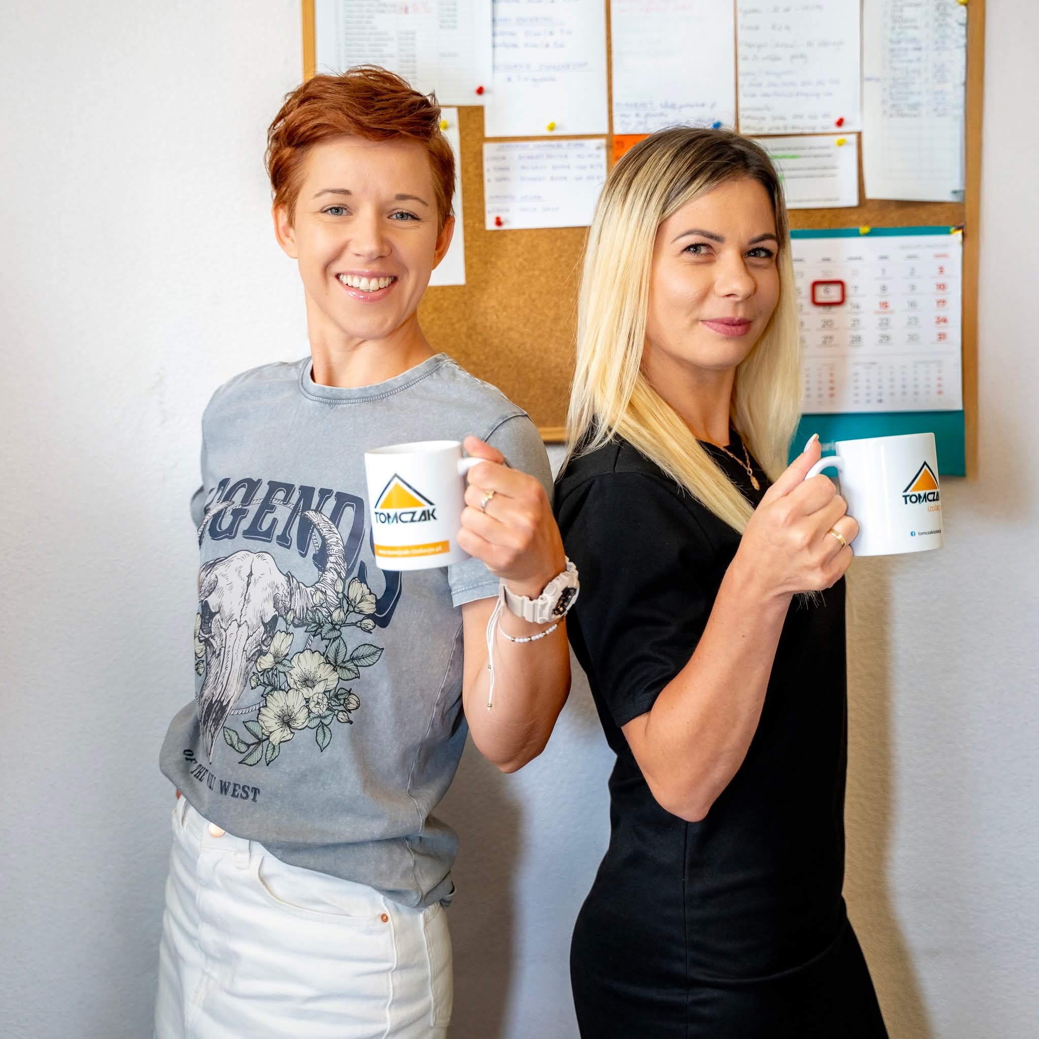 Dwie uśmiechnięte kobiety z kubkami firmy Tomczak Izolacje na tle tablicy korkowej z kalendarzem i notatkami.