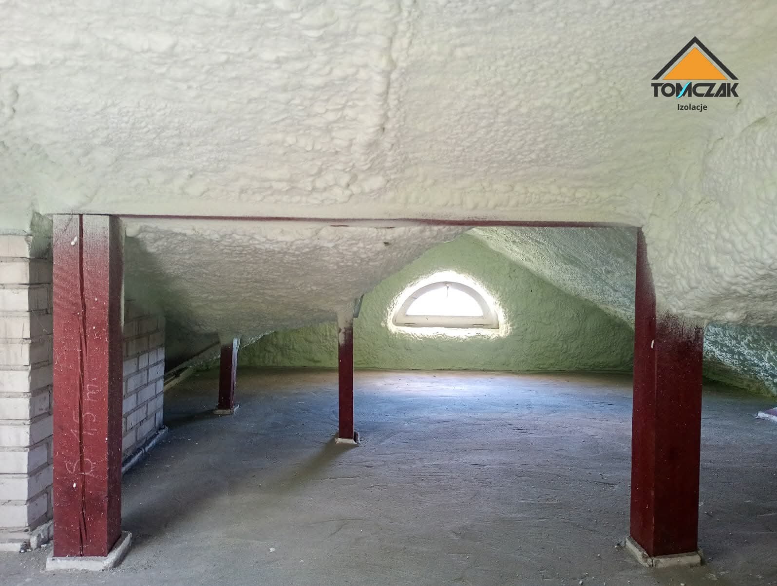 Poddasze z oknem po ociepleniu pianką PUR, widoczne drewniane słupy i logo firmy Tomczak Izolacje w prawym górnym rogu. Izolacja pokrywa skośne ściany i sufit.