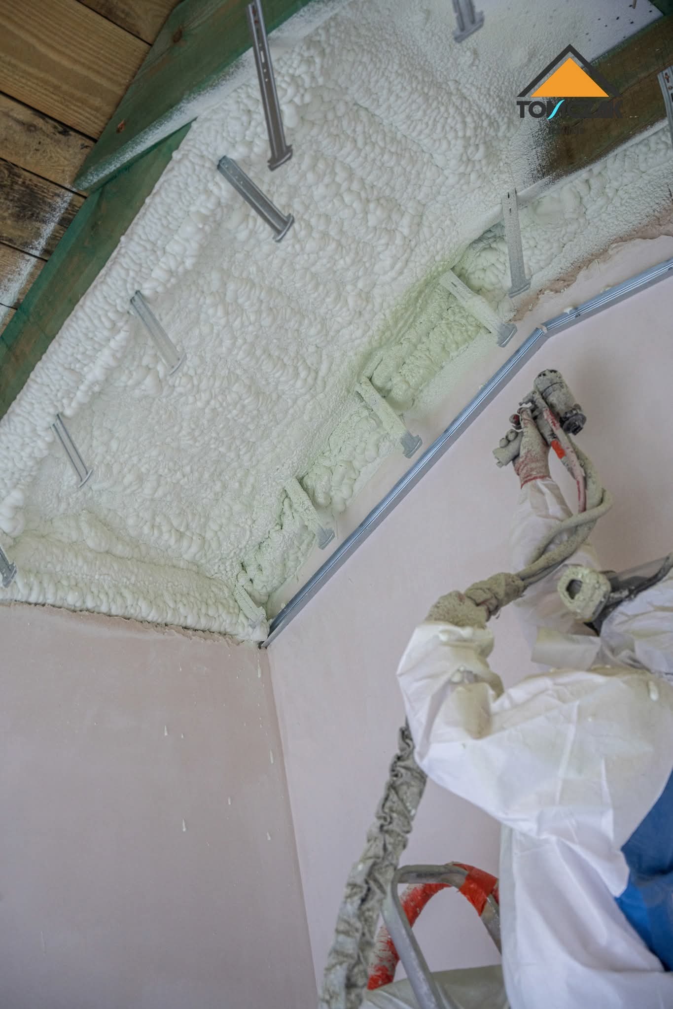 Poddasze ocieplone pianką PUR z widocznymi mocowaniami i aplikatorem. Praca w toku, pianka świeżo nałożona, widoczne detale struktury i sprzęt ochronny.