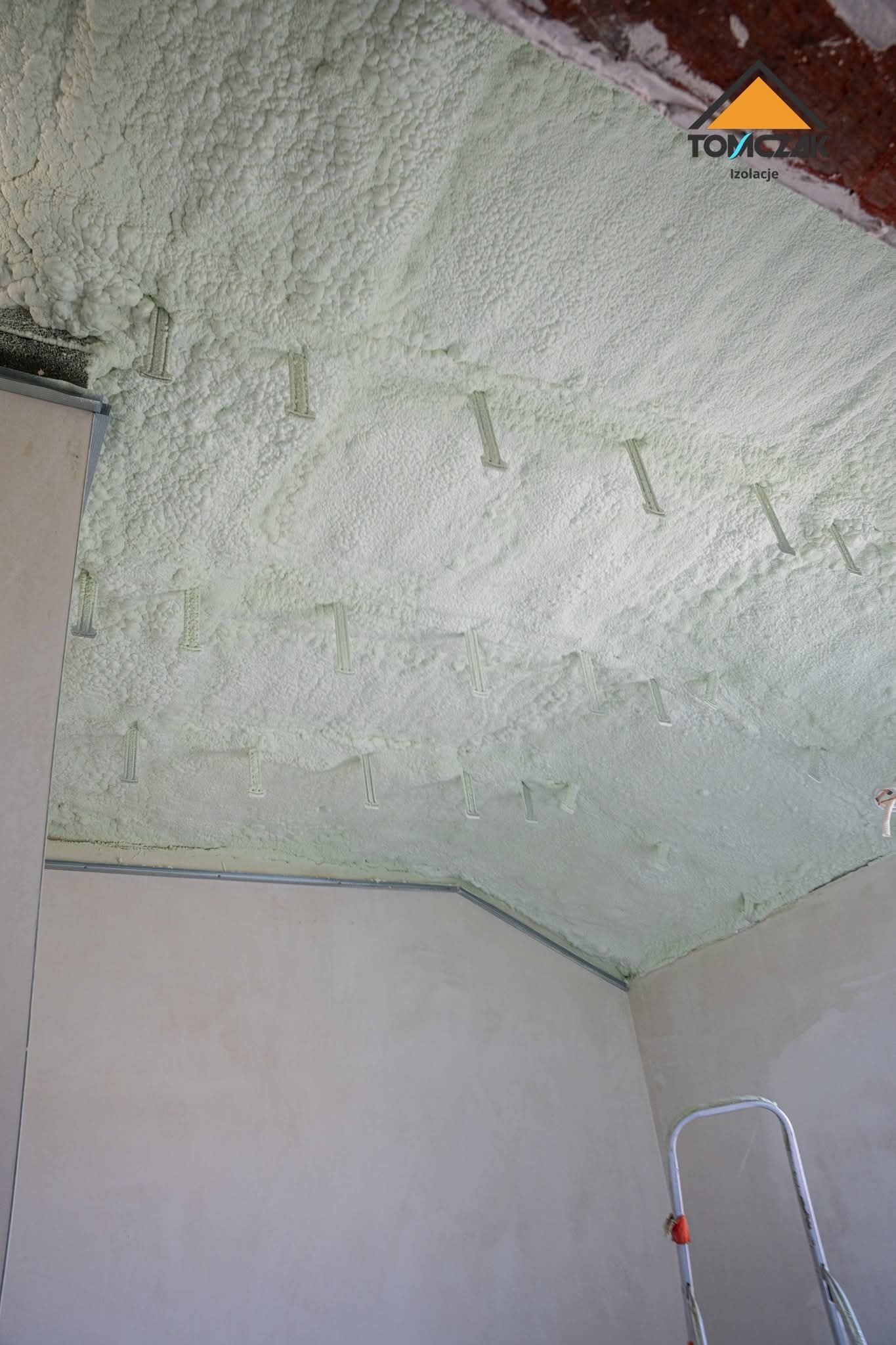 Poddasze po aplikacji pianki PUR, widoczne mocowania. Izolacja pokrywa całą powierzchnię, tworząc jednolitą warstwę. W tle ściana i drabina.