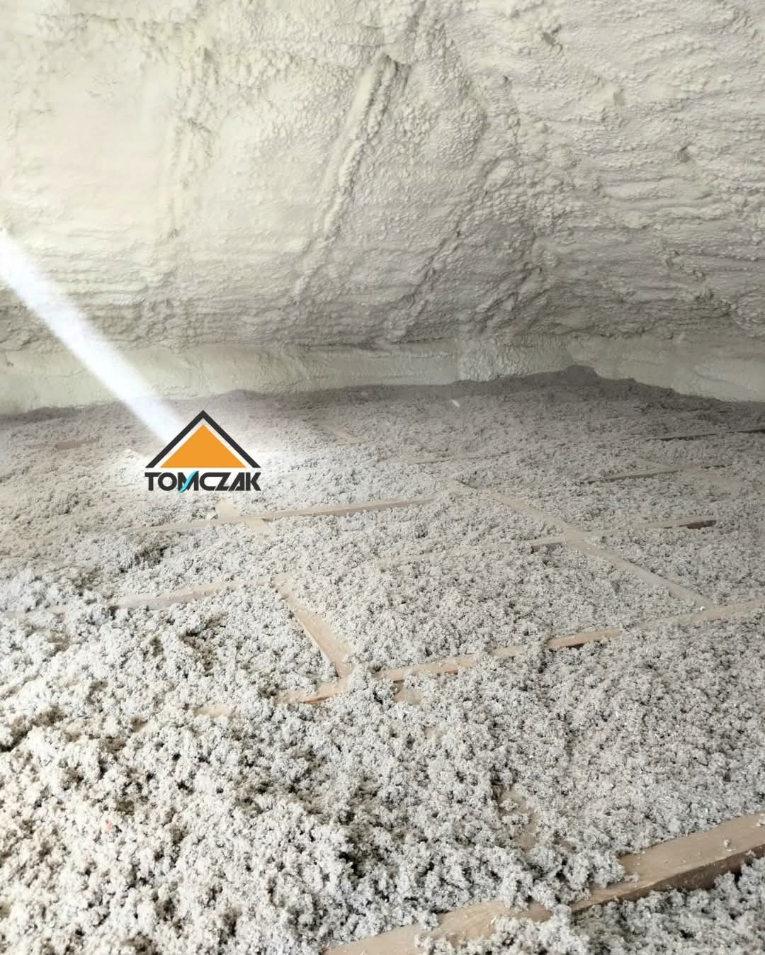 Poddasze po ociepleniu pianką PUR, widoczne drewniane belki konstrukcyjne, nierównomierna struktura pianki, logo firmy w lewym dolnym rogu.