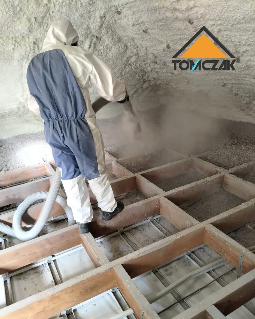 Pracownik w kombinezonie aplikuje pianę PUR na poddaszu. Widoczne drewniane legary i warstwa izolacji. Logo firmy w prawym górnym rogu.