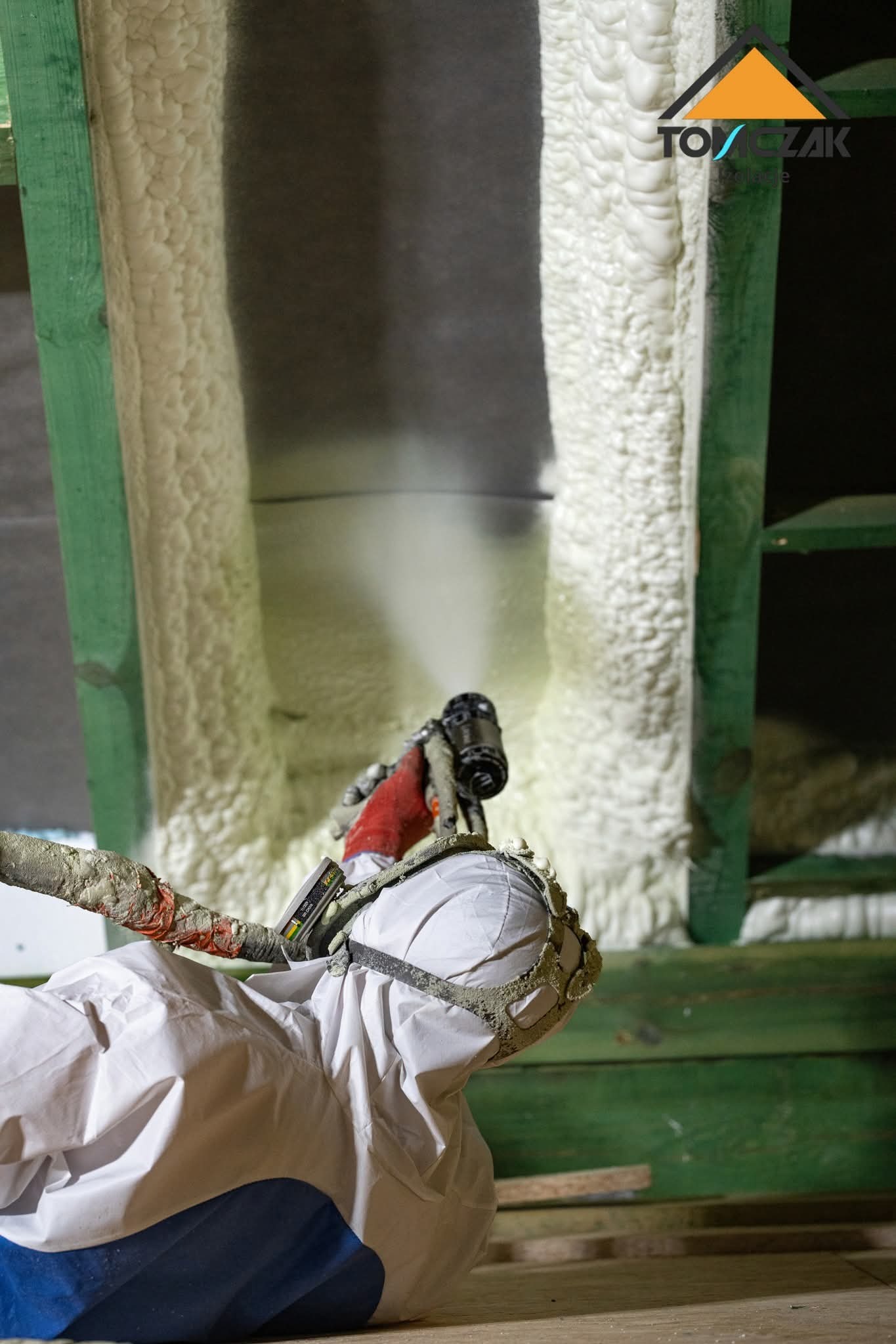 Pracownik w kombinezonie nanosi pianę PUR na drewnianą konstrukcję, tworząc izolację termiczną. Widoczny pistolet natryskowy i fragment ocieplonej powierzchni.