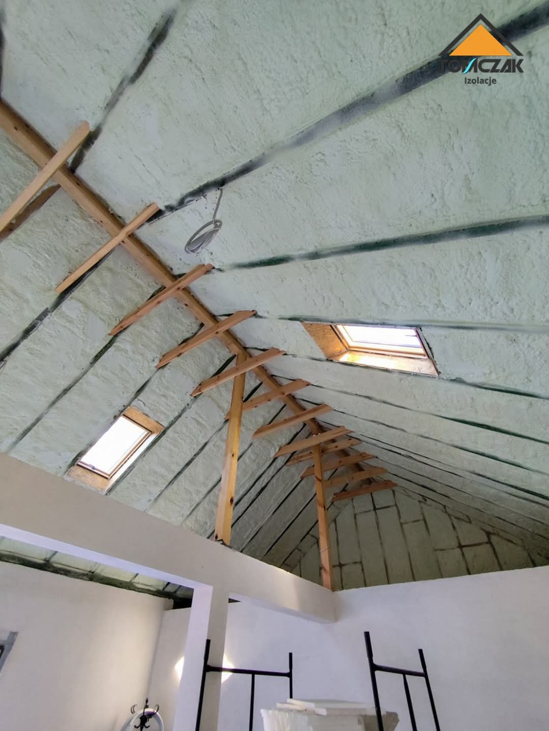 Poddasze po ociepleniu pianką PUR, widoczne drewniane elementy konstrukcyjne i skosy dachu. Dwa okna dachowe wpuszczają światło. Widok z dołu, z fragmentem pomieszczenia.