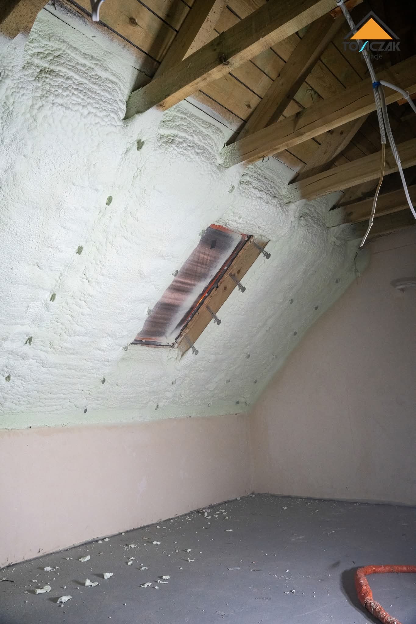 Poddasze po ociepleniu pianką PUR, widoczne krokwie i okno dachowe. Biała, nierówna powierzchnia pianki. Na podłodze resztki pianki, ściany pomalowane na jasny kolor.