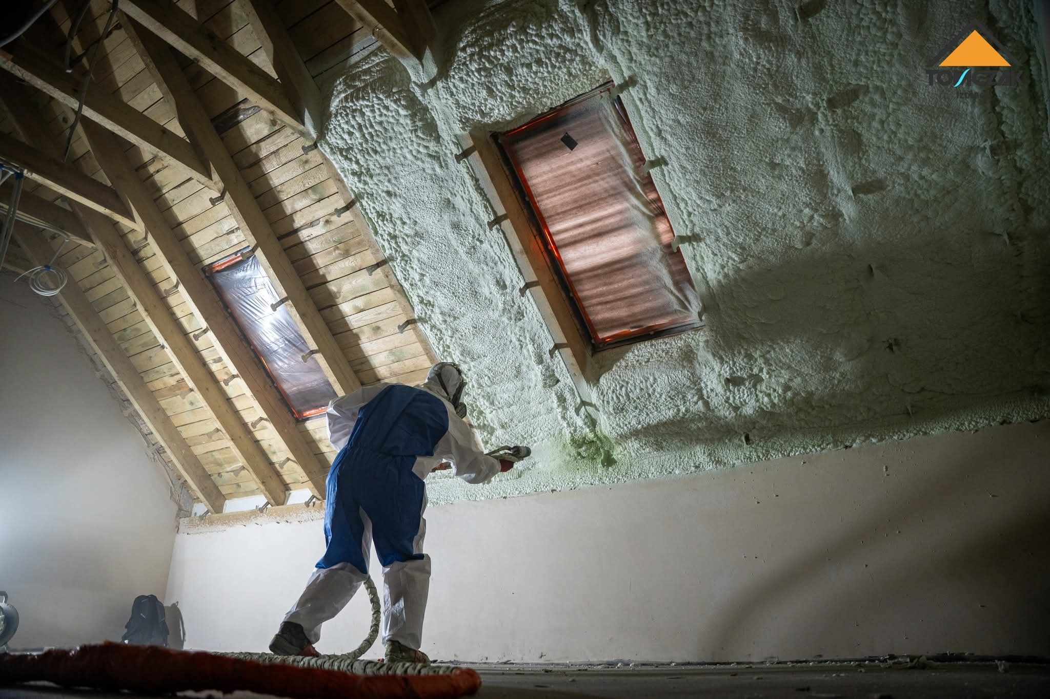 Pracownik w kombinezonie natryskuje pianę PUR na poddasze z oknami, widoczne drewniane krokwie i ocieplona ściana. Ocieplanie pianką PUR poddasza.