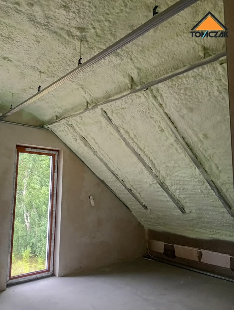 Poddasze po ociepleniu pianką PUR, widoczne metalowe profile konstrukcyjne, okno dachowe z widokiem na zieleń. Świeżo ocieplone poddasze z widoczną fakturą pianki.