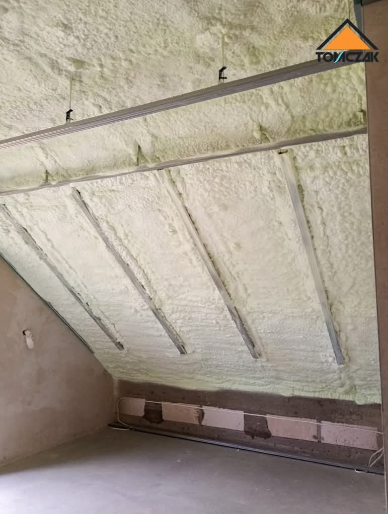 Poddasze ocieplone pianką PUR, widoczne metalowe profile montażowe. Ściana w stanie surowym, z widocznymi bloczkami i przewodami. Oświetlenie górne.