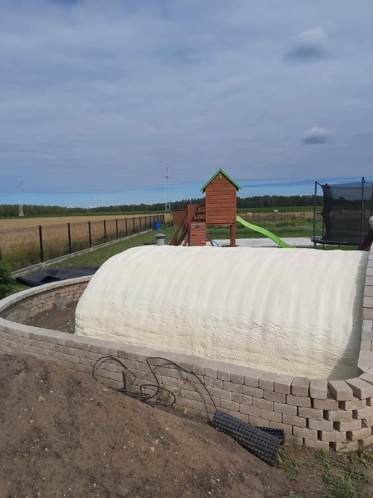 Kopuła pokryta białą pianą PUR, otoczona murkiem z kostki brukowej, na tle placu zabaw i krajobrazu polnego. Ocieplenie pianką poliuretanową.
