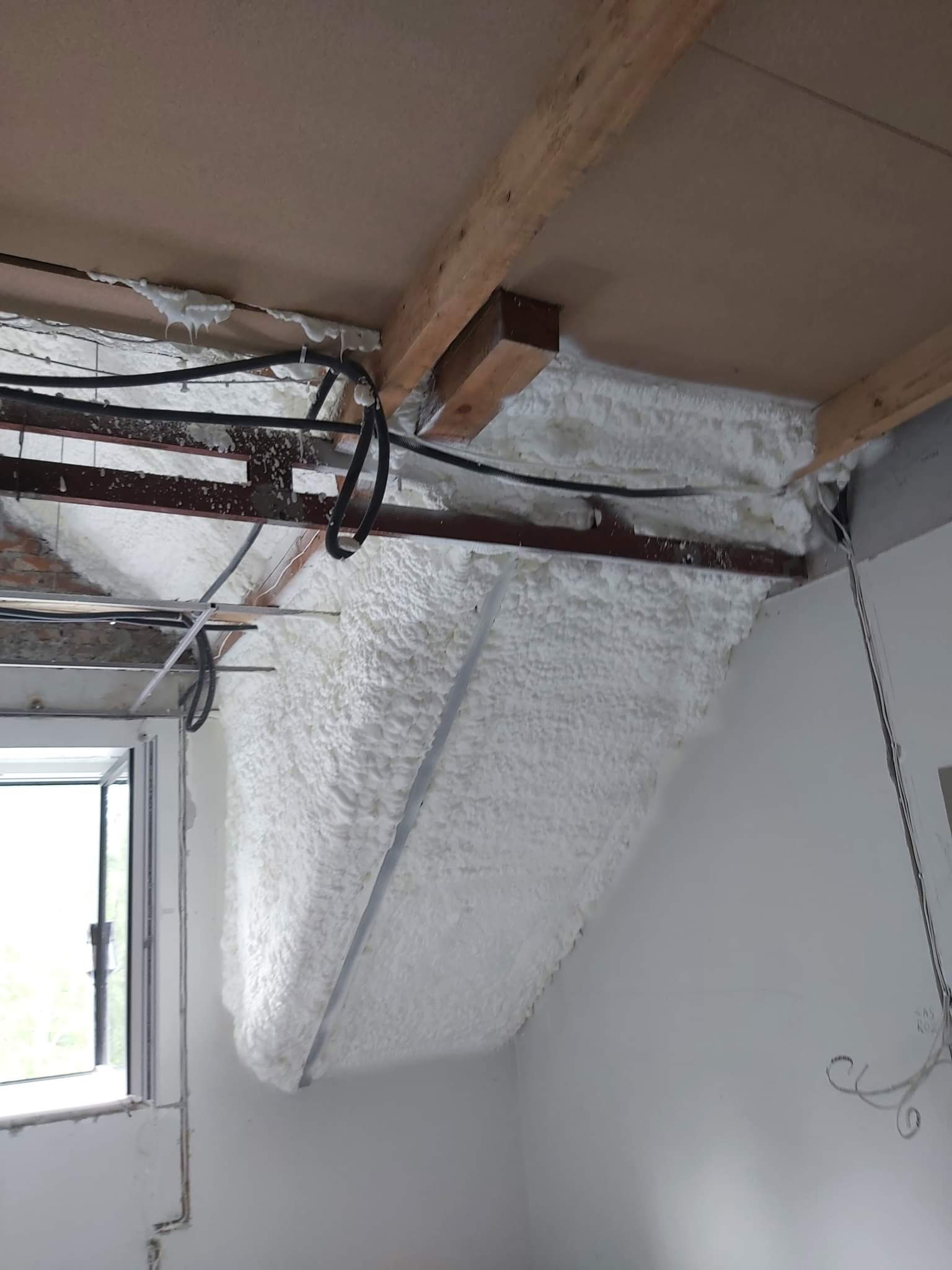 Poddasze z widoczną białą pianką izolacyjną, aplikowaną między krokwiami i pod stropem. Widoczne przewody elektryczne i fragment okna.