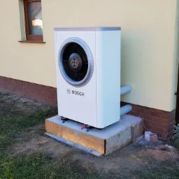 Biała pompa ciepła Bosch zamontowana na betonowym fundamencie przy ścianie domu z ceglaną podmurówką i brązową rynną.