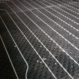 Białe rury ogrzewania podłogowego ułożone na czarnej, profilowanej macie izolacyjnej, widok z góry pod kątem, fragment płyty OSB w prawym górnym rogu.