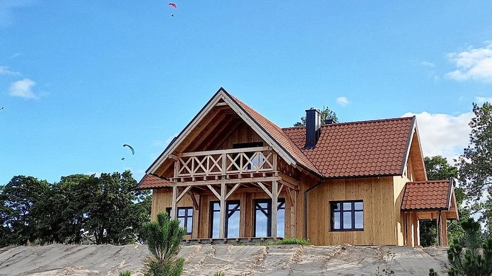 Jednorodzinny dom murowany w stanie surowym otwartym z drewnianą konstrukcją balkonu, czerwoną dachówką ceramiczną i czarnymi ramami okiennymi na tle błękitnego nieba z paralotniarzami.