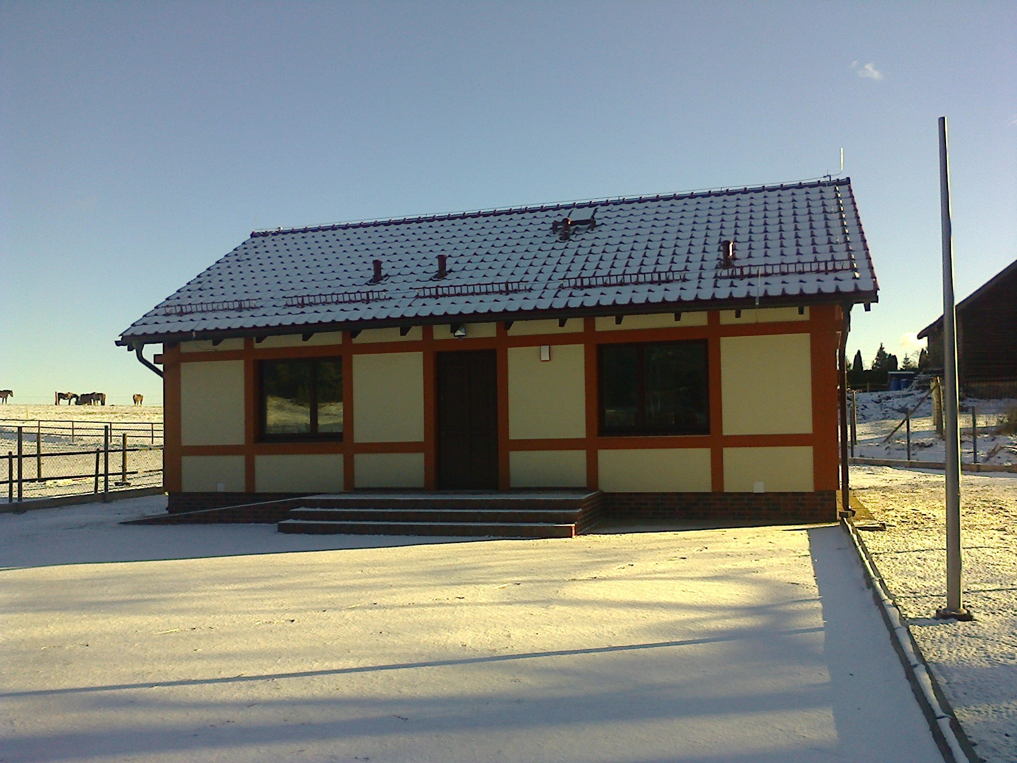 Leśniczówka w stylu muru pruskiego z ceramiczną dachówką, częściowo pokryta śniegiem, na tle zaśnieżonego krajobrazu z widocznymi końmi na pastwisku.