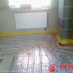 Białe rury ogrzewania podłogowego ułożone na siatce z czerwonymi liniami, przygotowane do zalania wylewką, widoczny grzejnik na ścianie i fragment kolumny w rogu pomieszczenia.