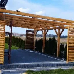 Drewniana pergola z lamelami i widocznym szkieletem dachu, częściowo zadaszona kostką brukową, z drabinką i widokiem na pola uprawne w tle.