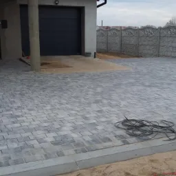 Ułożona szara kostka brukowa na podjeździe przed domem z garażem, widoczny kabel elektryczny na piasku przy krawężniku.