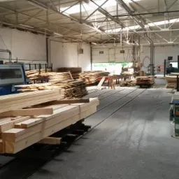 Wnętrze hali produkcyjnej z ułożonymi stosami świeżo obrobionego drewna, widoczna maszyna do obróbki drewna z systemem odsysania pyłu oraz pracownicy w tle