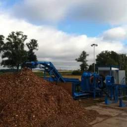 Duża pryzma zrębków drzewnych przed niebieską linią produkcyjną i budką operatora w tle, widok w pochmurny dzień.