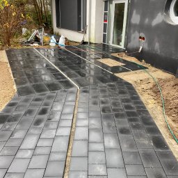 ALL-BRUK Alan Grzyb - Układanie ciemnoszarej kostki betonowej na posesji, widoczne przerwy między kostkami wypełnione drobnym żwirem, dom w tle.