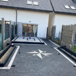 Ułożona ciemnoszara nawierzchnia z wzorem kompasu z jasnych elementów, obrzeża z czarnej kostki, w tle budynek z szarą elewacją i ciemnym dachem.