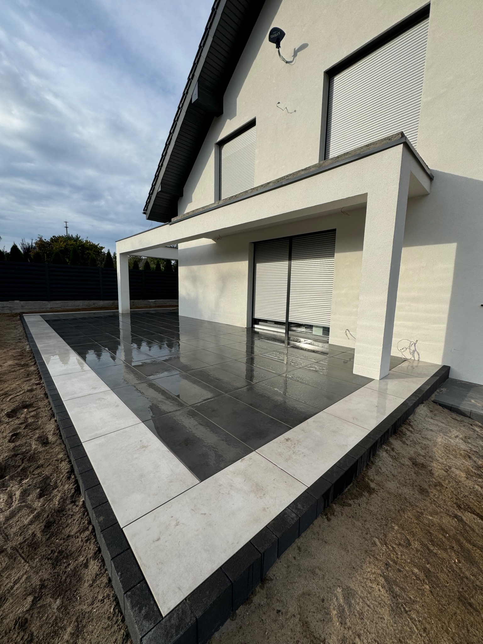 Nowoczesny taras z ciemnych płyt wykończony jasnymi płytami i obrzeżem z kostki, przylegający do białego domu z szarymi roletami.