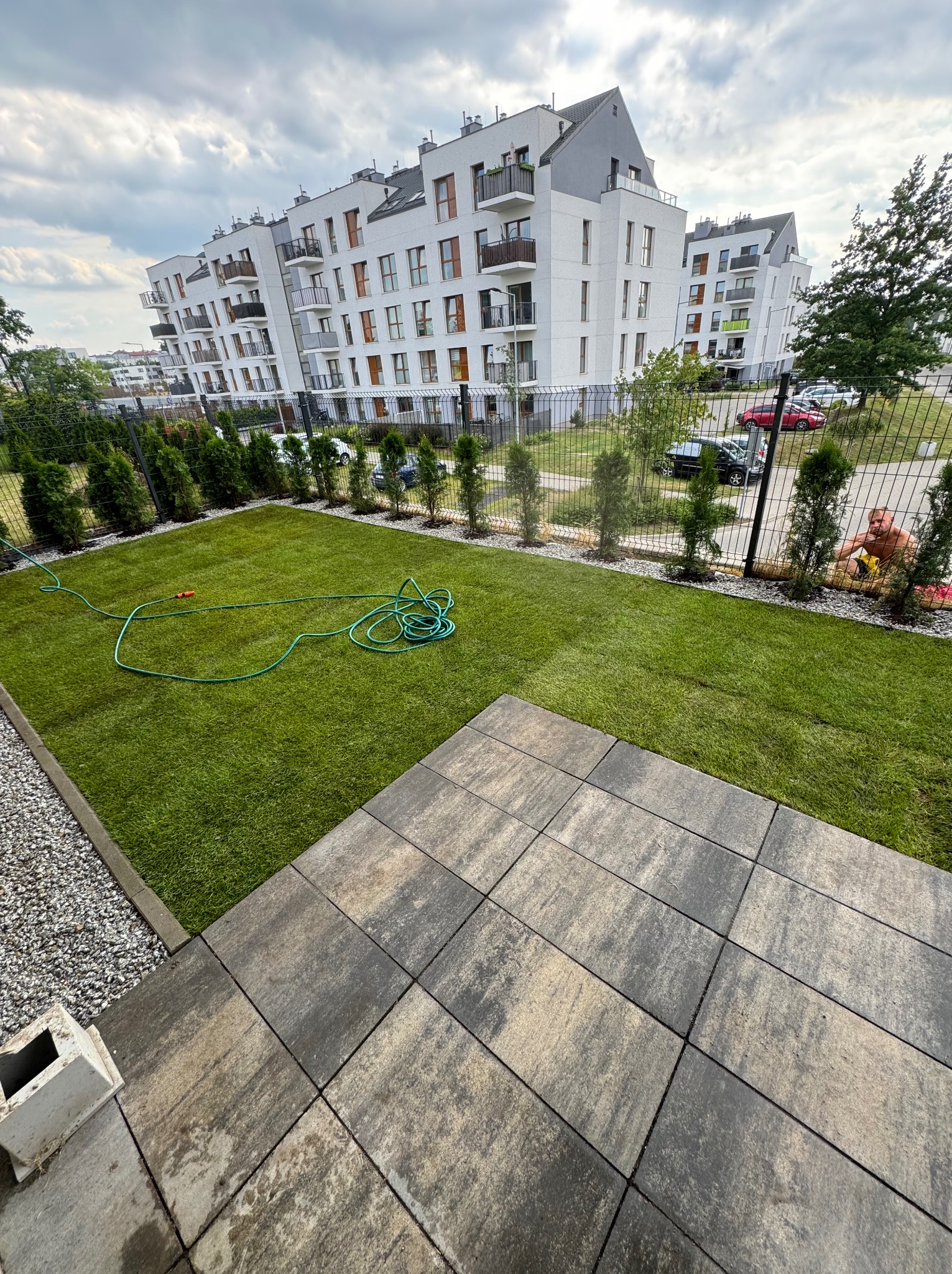 Ułożona nawierzchnia z szarych płyt na tle trawnika z systemem nawadniania i zabudowy wielorodzinnej w tle.