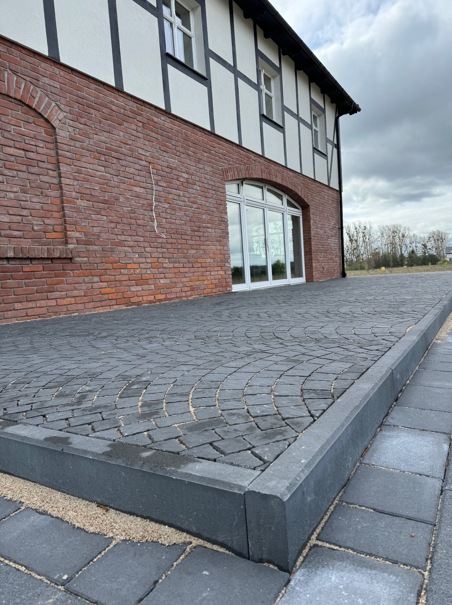 Ułożona nawierzchnia z ciemnoszarej kostki brukowej w kształcie wachlarza przy budynku z czerwonej cegły i białej fasady szachulcowej, widoczne obrzeża i piasek między kostkami.