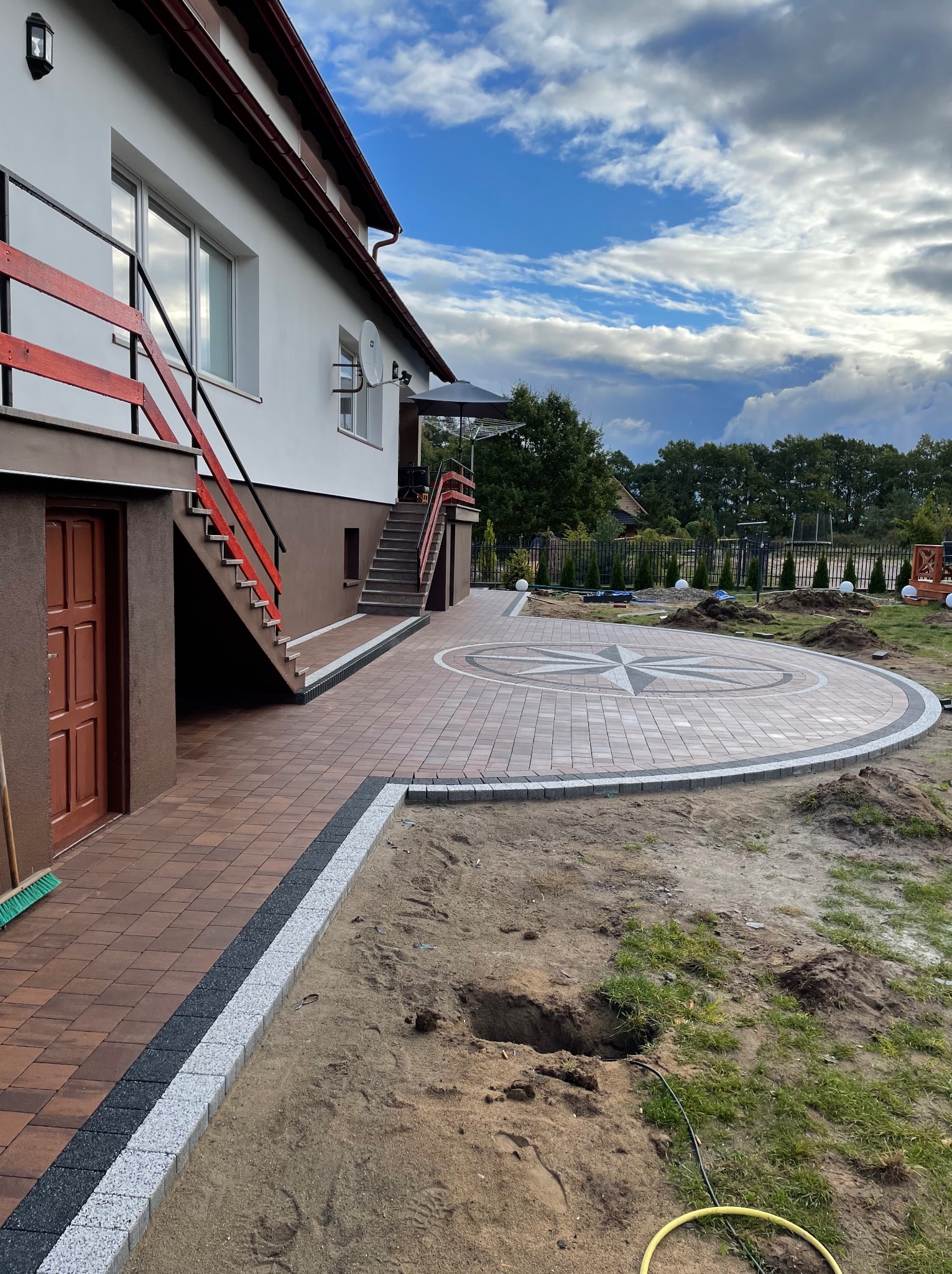 Układanie nawierzchni z kostki brukowej w kształcie kompasu na posesji, widoczne niedokończone obrzeża i wykopane doły w ziemi, w tle dom z czerwonymi barierkami i zielony ogród.