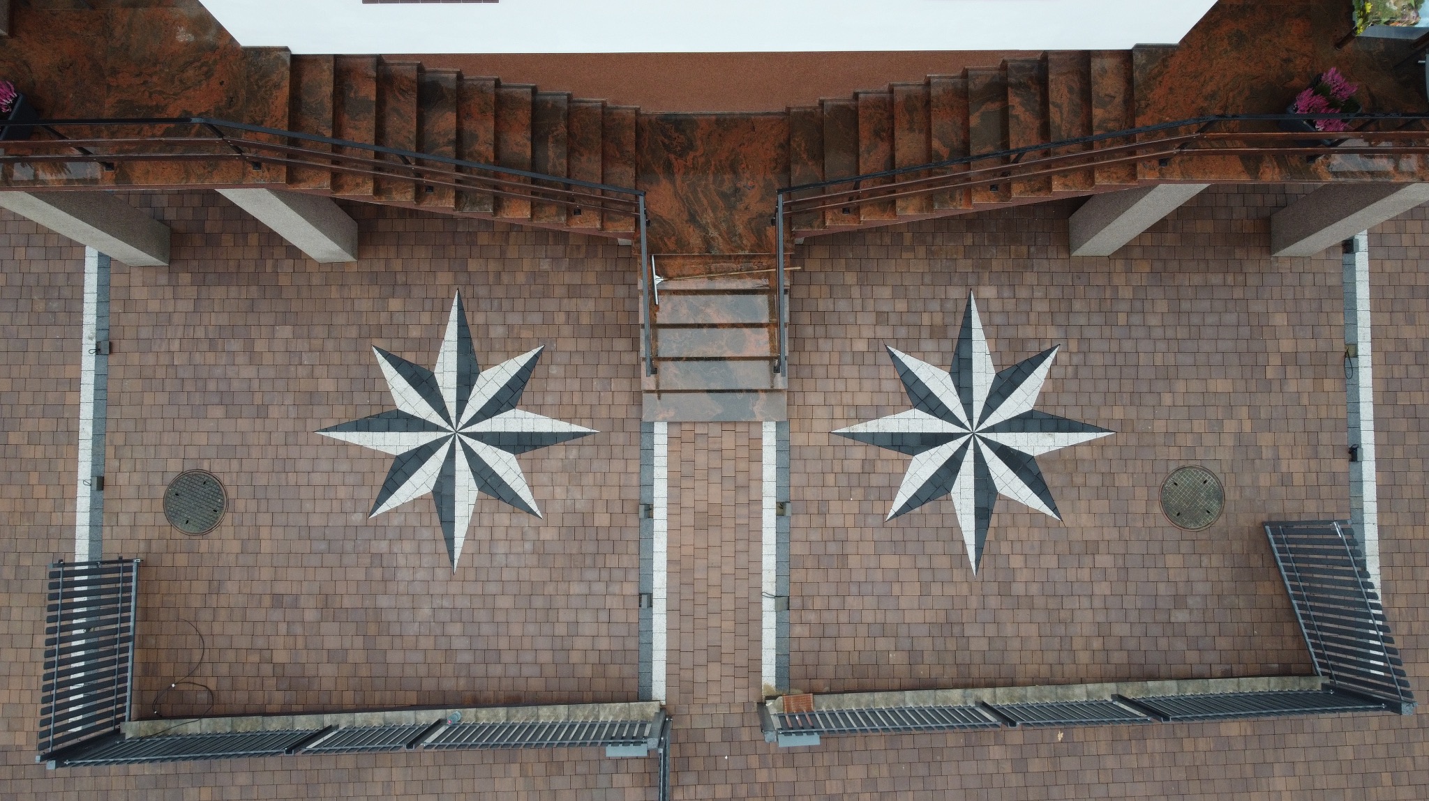 Widok z góry na dziedziniec z brązowej nawierzchni z wzorem gwiazdy, schody z czerwonego granitu i elementy małej architektury.