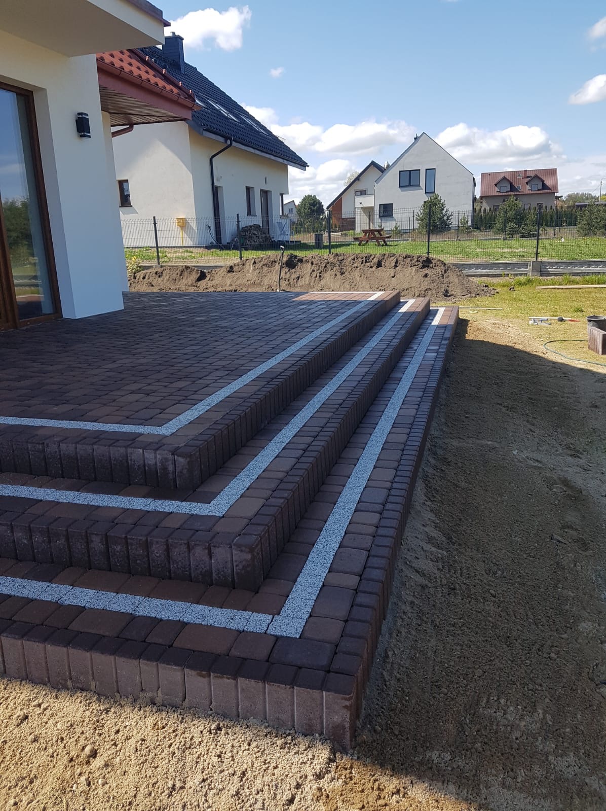 Ułożone schody z ciemnobrązowej kostki z jasnymi fugami, prowadzące z tarasu do ogrodu, w tle domy jednorodzinne w słoneczny dzień.