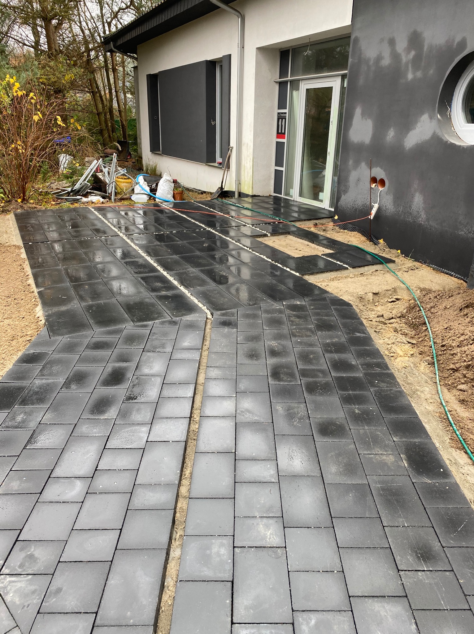 Układanie ciemnoszarej kostki betonowej na posesji, widoczne przerwy między kostkami wypełnione drobnym żwirem, dom w tle.