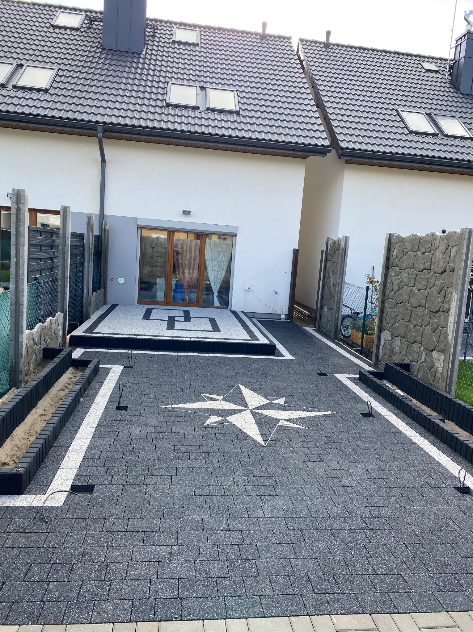Ułożona ciemnoszara nawierzchnia z wzorem kompasu z jasnych elementów, obrzeża z czarnej kostki, w tle budynek z szarą elewacją i ciemnym dachem.